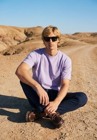 Jeune homme aux cheveux blonds portant des lunettes de soleil, un t-shirt violet, un pantalon foncé et des chaussures marron, assis les jambes croisées sur une route en terre dans un paysage sec et rocailleux.