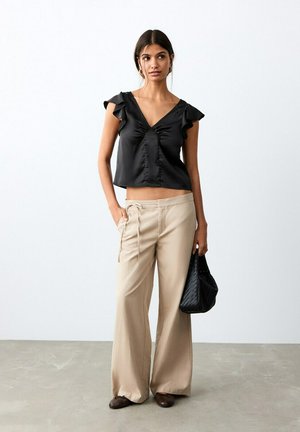 Žena oblečená v čiernom top s volánovými rukávmi, béžové nohavice s širokými nohavicami, hnedé topánky, drží čiernu textúrovanú kabelku, stojí na sivom podlahe.