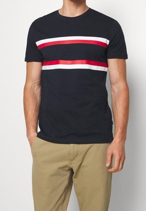 Homme portant un t-shirt noir à manches courtes avec deux bandes horizontales rouges et blanches sur la poitrine, associé à un pantalon beige.