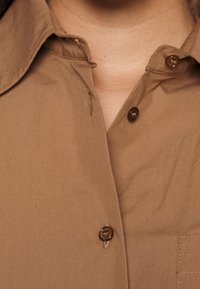 Camicia button-up beige chiaro con colletto, caratterizzata da bottoni marroni, texture liscia e un piccolo taschino sul davanti.
