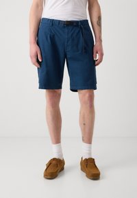Shorts bleu marine avec une taille ceinturée, deux poches latérales et une coupe décontractée. Portés avec des chaussures en daim marron et des chaussettes blanches.