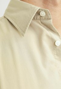 Beige Hemd mit Knopfleiste und spitzen Kragen. Aus glattem Stoff gefertigt, mit weißen Knöpfen und dezenten Nähedetails am Kragen.