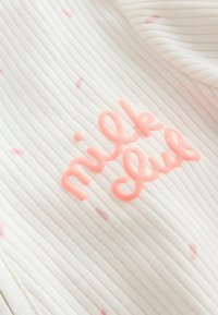 Tissu blanc côtelé avec de petits pois roses et un texte en relief rose en écriture cursive disant "milk club" à la surface.