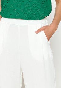 Weiße, leichte Hosen mit Seitentaschen und einer glatten Textur, kombiniert mit einem grünen Oberteil mit Ösenmuster in lockerer Passform.