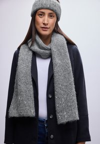 Sciarpa grigia in maglia con un motivo strutturato e paillettes argentate, abbinata a una fascia per capelli grigia coordinata, indossata sopra un cappotto nero e una maglietta bianca.