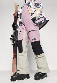 Skiantrekk bestående av en rosa magebelte med svarte og beige detaljer, kombinert med en mønstret jakke og svarte skistøvler. Skiutstyr inkludert.