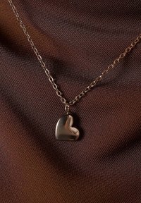 Zilveren hart hangerketting op een delicate ketting tegen een gestructureerde bruine stoffen achtergrond. Glad oppervlak, ronde randen en gepolijste afwerking.
