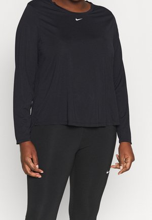 Osoba ubrana w czarną, długorękawową koszulkę Nike i czarne legginsy Nike, stoi z rozluźnionymi rękami wzdłuż ciała.