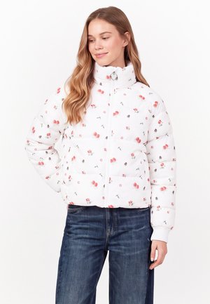 Frau mit langen Haaren, die eine weiße Daunenjacke mit kleinen roten Blumenmustern und blaue Jeans trägt, steht vor einem schlichten weißen Hintergrund.