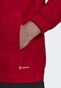 Rød jakke med elastiske manchetter og sidelommer, lavet af letvægtsmateriale. Har et "AEROREADY" logo nederst på kanten.