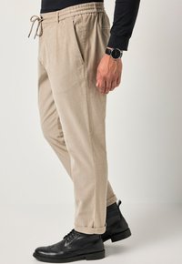 Pantaloni in corduroy beige con una vestibilità rilassata, vita con cordoncino, tasche laterali e stivali neri con lacci. Design con frontale piatto e gamba affusolata.
