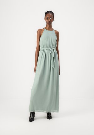 Robe maxi vert clair avec un col halter et une ceinture à la taille, confectionnée dans un tissu lisse, silhouette fluide, associée à des bottes noires à la cheville.