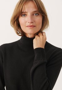 Črni pulover s turtleneck iz mehkega pletenega materiala, ki ima rebraste manšete in visok ovratnik. Minimalističen dizajn in gladka tekstura.