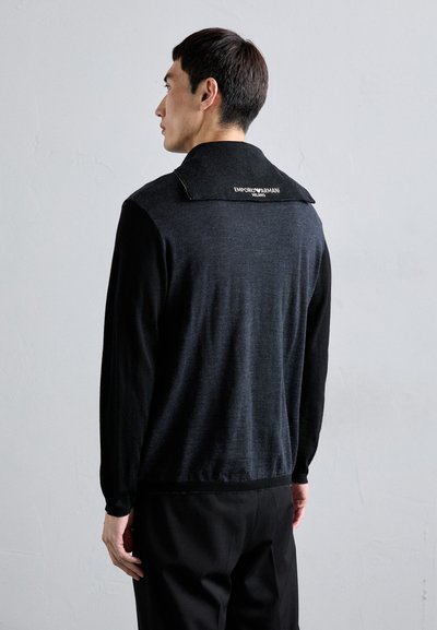 A balra néző férfi fekete és sötétszürke hosszú ujjú pulóvert visel, amelynek a vállán egy "Emporio Armani Milano" feliratozott lap található.