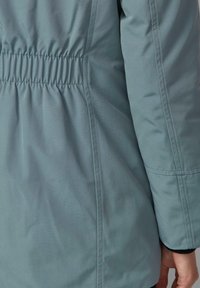 Close-up osobe koja nosi svijetloplavu jaknu s nabranim elastičnim pojasom i dugim rukavima, ruka opuštena uz bok.