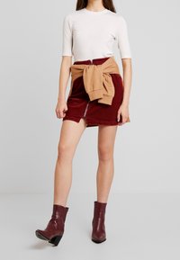 Bílý top s krátkým rukávem, zkombinovaný s vínově červenou mini sukní z manšestru, která má přední zip, a béžovou mikinou uvázanou v pase. Hnědé kotníkové boty.