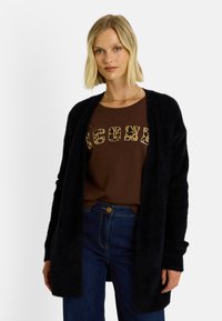 Cardigan noir et duveteux avec un design à ouverture frontale, porté sur un t-shirt marron avec des lettres à imprimé léopard, associé à un jean bleu.