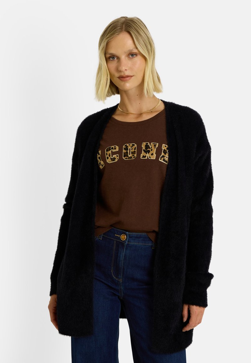 Cardigan noir et duveteux avec un design à ouverture frontale, porté sur un t-shirt marron avec des lettres à imprimé léopard, associé à un jean bleu.