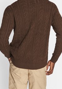 Brązowy sweter z dzianiny, z wzorem warkoczy, ściągaczami przy rękawach i u dołu. Widok z tyłu ukazuje teksturowany design i dopasowanie.