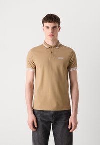 Tansfarbenes Polohemd mit Kragen, das abwechselnd weiße und beige Streifen aufweist, kurze Ärmel und ein Logo-Patch auf der Brust hat. Getragen mit dunklen Jeans.