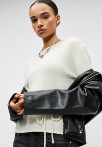Femme aux cheveux plaqués en arrière portant un pull blanc, un collier en grosse chaîne argentée, et une veste en cuir noir posée sur ses bras.