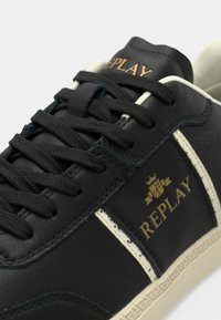 Baskets en cuir noir avec des accents blancs et un bout texturé. Dotées de lacets noirs et d'un logo doré sur le côté.