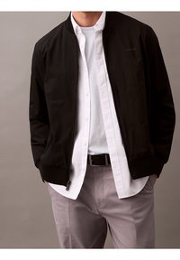 Schwarze Bomberjacke mit Reißverschluss, gerippten Bündchen und Saum, kombiniert mit einem weißen Hemd mit Knöpfen. Graue Hose vervollständigt das Outfit.