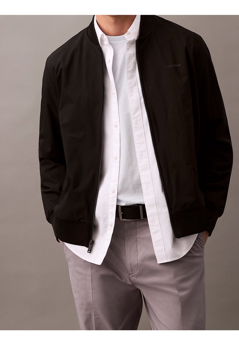 Schwarze Bomberjacke mit Reißverschluss, gerippten Bündchen und Saum, kombiniert mit einem weißen Hemd mit Knöpfen. Graue Hose vervollständigt das Outfit.