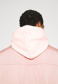 Chaqueta con capucha rosa, con un suave cuello elevado y un tejido exterior liso y brillante; presenta un diseño relajado y costuras visibles en la parte posterior.