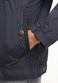 Marineblauwe jas met een gladde textuur, voorzien van een zijvak, knoopaccenten en contrasterende stiksels langs de mouw en de zoom.