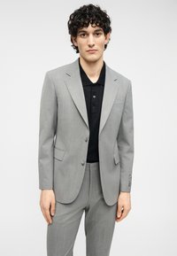 Γκρι κοστούμι κατά παραγγελία με σακάκι μονόπετο και εφαρμοστά παντελόνια. Κάτω από αυτό ένα μαύρο πόλο πουκάμισο. Το ύφασμα έχει μια διακριτική υφή.