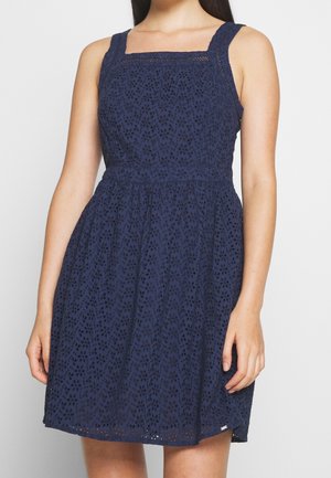 Woman wearing a sleeveless navy blue eyelet dress with a square neckline and fitted waist against a plain background.