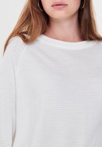 Primo piano di una giovane donna con lunghi capelli castano chiaro, che indossa orecchini a cerchio in oro e un maglione bianco a maglia testurizzata.