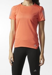 Kobieta ubrana w koralową sportową koszulkę z krótkim rękawem oraz czarne legginsy, obie części odzieży z logo Adidas, stojąca z rozluźnionymi rękami przy bokach.