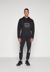 Felpa nera con cappuccio e testo bianco, abbinata a pantaloni della tuta neri con strisce bianche sulle fasce laterali. Sneakers nere ai piedi.