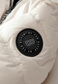 Puffermantel in Creme mit einem runden schwarzen Logopatch. Verfügt über eine gesteppte Textur und schwarze Reißverschlüsse, was ein modernes, sportliches Design unterstreicht.