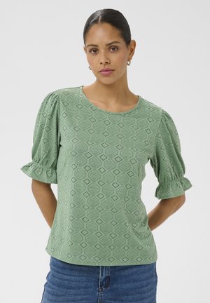 Žena v svetlozelenej blúzke s potlačou, s nafúknutými krátkymi rukávmi a modrými džínmi, stojaca pred jednoduchým svetlým pozadím.