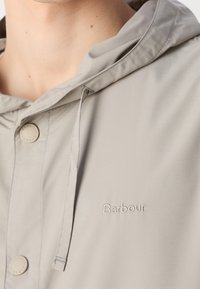 Zbliżenie jasnoszarej kurtki z kapturem z haftowanym logo Barbour oraz zatrzaskami na klatce piersiowej i sznurkiem wiszącym w dół.