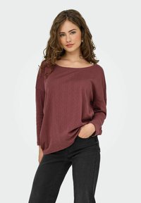 Maglione borgogna con texture, scollatura a barca, spalle scese e maniche lunghe, abbinato a jeans neri.