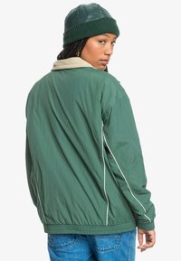 Zelená nylónová bunda s fleece podšitým golierom, elastickými manžetami a bielymi kontrastnými prúžkami. Nosená so zelenou pletenou čiapkou.
