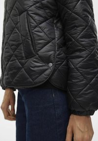 Černá prošívaná bunda s diamantovým vzorem, postranní kapsou na knoflík a elastickými manžetami, kombinovaná s tmavými džínami.