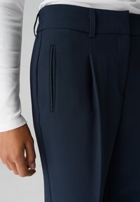 Pantaloni blu navy con una texture liscia, dotati di fronte piatto, tasche laterali e una vestibilità su misura. Mano appoggiata sul girovita.