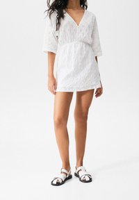 Weißes Kleid aus Spitzenstoff mit V-Ausschnitt, dreiviertel Ärmeln und tailliertem Schnitt. Kombiniert mit weißen und schwarzen Riemchensandalen.