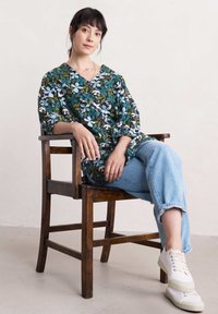 Tunika w kwiatowy wzór z dekoltem w serek, wykonana z tkaniny w odcieniach zieleni, niebieskiego i bieli. Połączona z jasnoniebieskimi dżinsami i białymi trampkami.