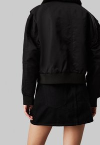 Veste bomber noire avec un col en fausse fourrure douce, des poignets et un ourlet côtelés, et une texture lisse et brillante. Associée à une jupe en denim noire.