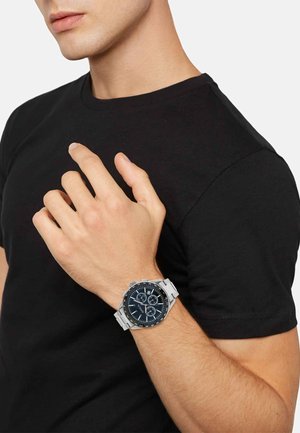 Sølv kronograf ur med en mørkeblå urskive, der har hvide markeringer og undermarkeringer, båret på en hånd mod en ensfarvet sort skjorte.