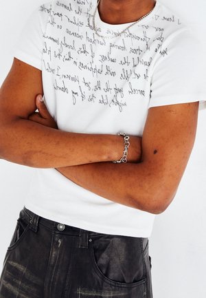 Witte kortekraag shirt met zwart handgeschreven tekst, gecombineerd met donkere, gestructureerde broek en een zilveren kettingarmband.