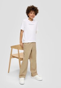 Weißes T-Shirt mit kurzen Ärmeln und aufgedrucktem Text, beige, locker sitzende Hose und weiße Sneaker. Holzstuhl im Hintergrund.