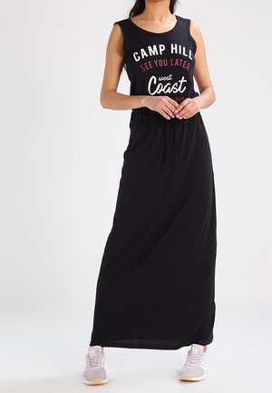 Frau trägt ein schwarzes ärmelloses Maxikleid mit dem Text "Camp Hill See You Later West Coast", kombiniert mit hellrosa Turnschuhen.