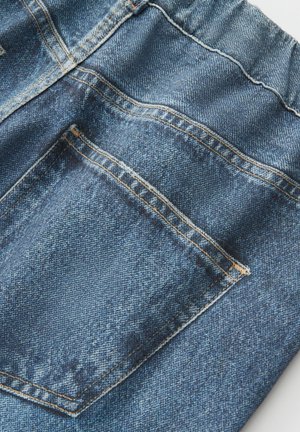 Close-up van blauwe spijkerbroek met zichtbare achterzak, oranje stiksel en details van de stoftextuur.